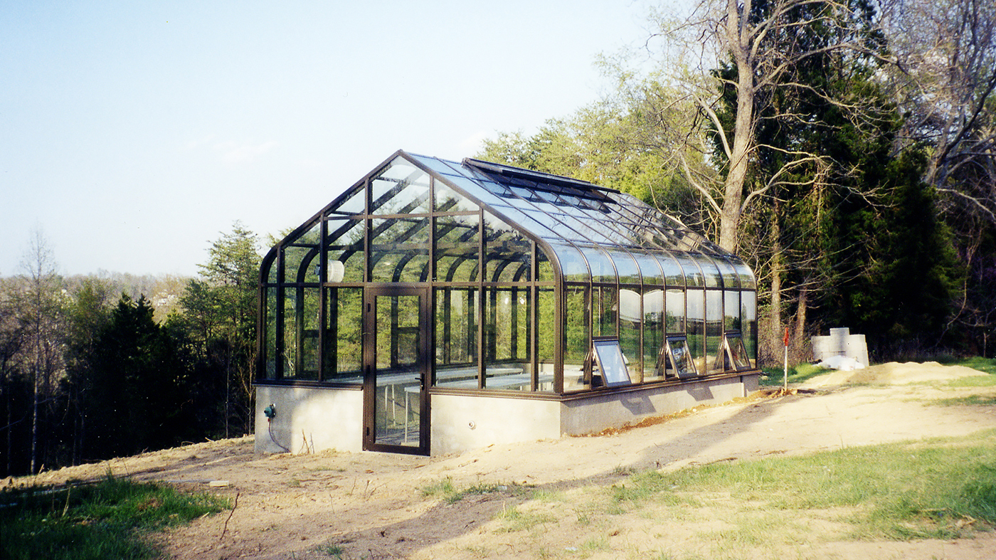 Curved eave, double pitch greenhouse with operable ridge vents, awning windows, terrace door. The interior of the greenhouse features a structural truss system, fixed benches with metal mesh tops, a humdifier and circulation fans.