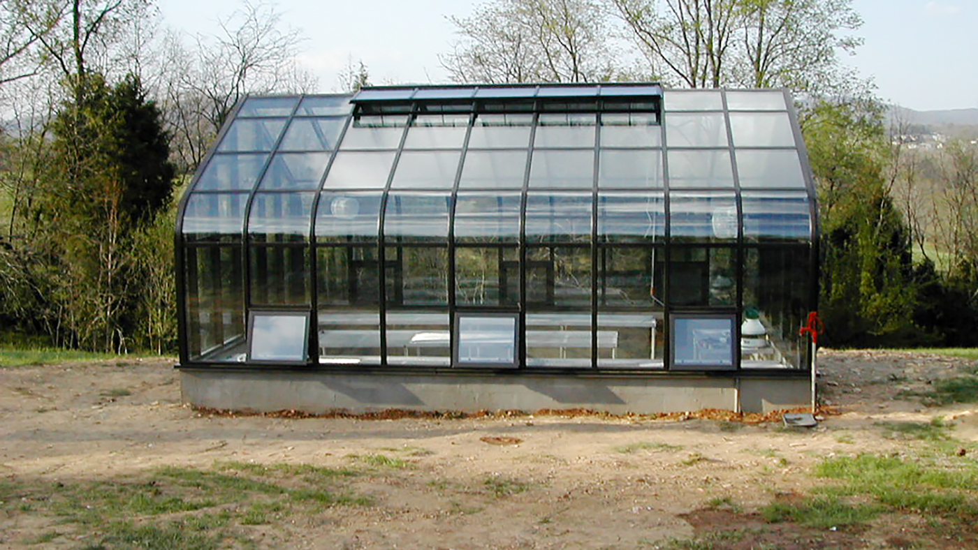 Curved eave, double pitch greenhouse with operable ridge vents, awning windows, terrace door. The interior of the greenhouse features a structural truss system, fixed benches with metal mesh tops, a humdifier and circulation fans.
