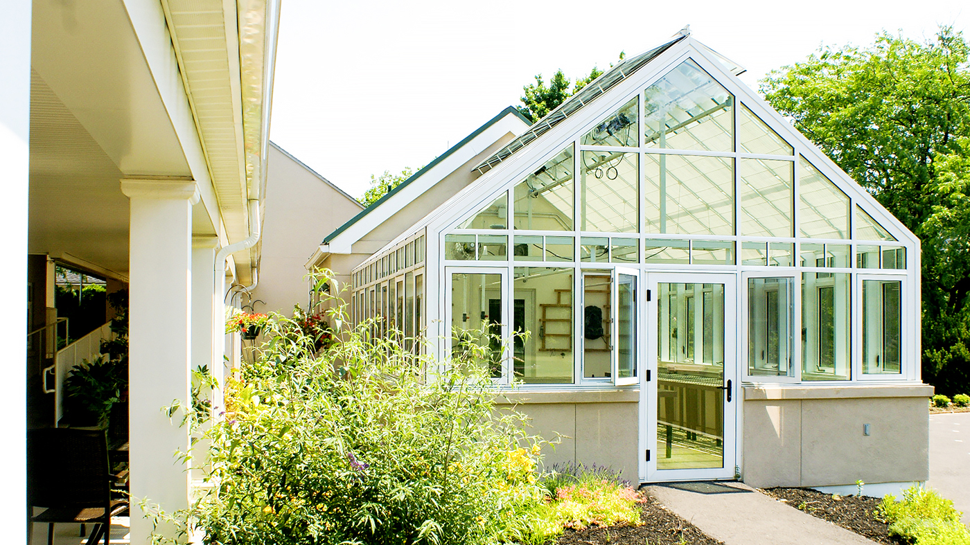 Straight eave double pitch greenhouse used at a retirement community. Unit includes ridge vents, transoms with grids, casement windows, terrace door, interior circulation fans, grow lights and interior roof mounted shades.